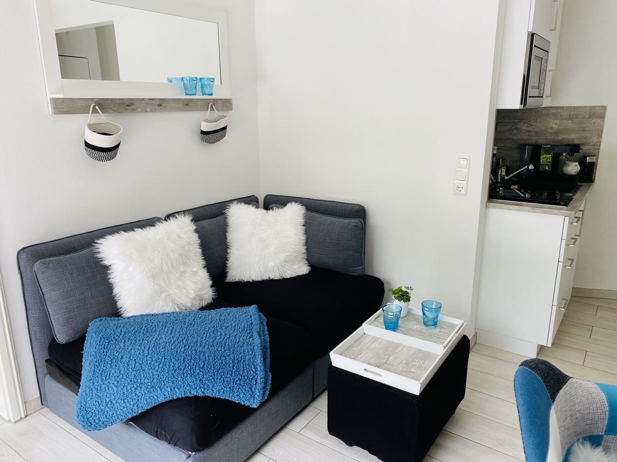 Schwarzes Sofa mit weißen Kissen und blauer Decke. Koffer-Tisch und Küchenbereich sichtbar.