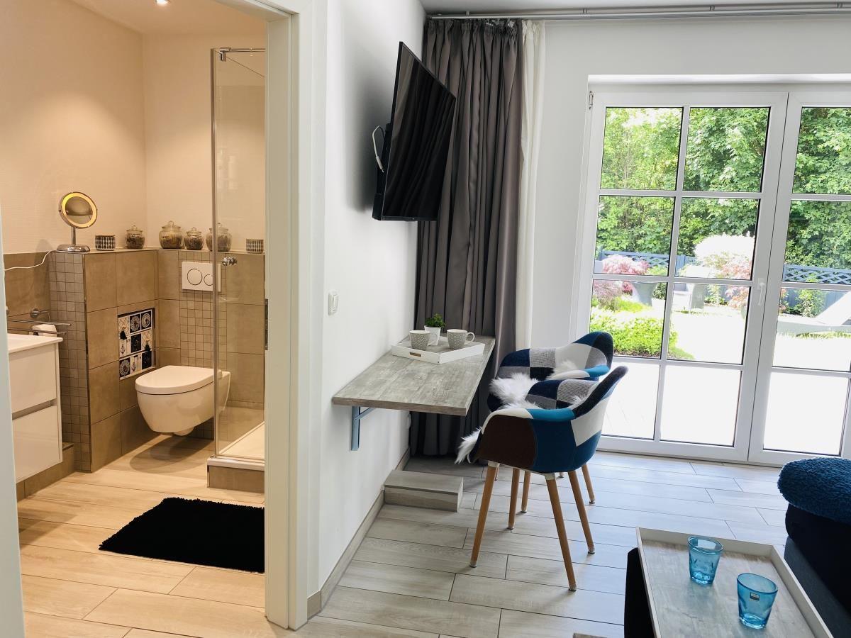 Wohnbereich mit TV, Schreibtisch und Blick auf Garten. Nebenraum mit Dusche und Toilette.
