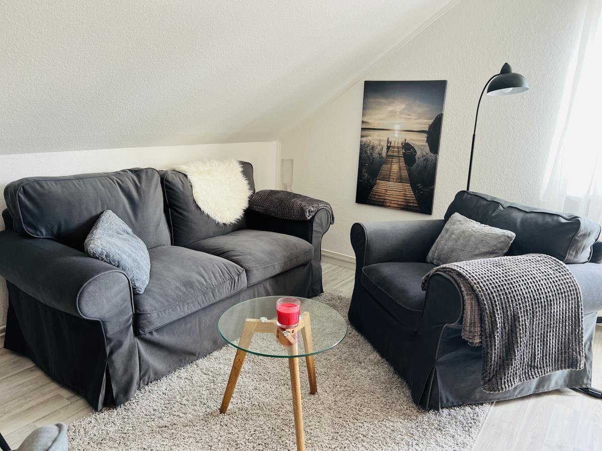 Wohnzimmer mit grauem Sofa, Sessel und Glas-Tisch. Wandbild und Stehlampe sind sichtbar.
