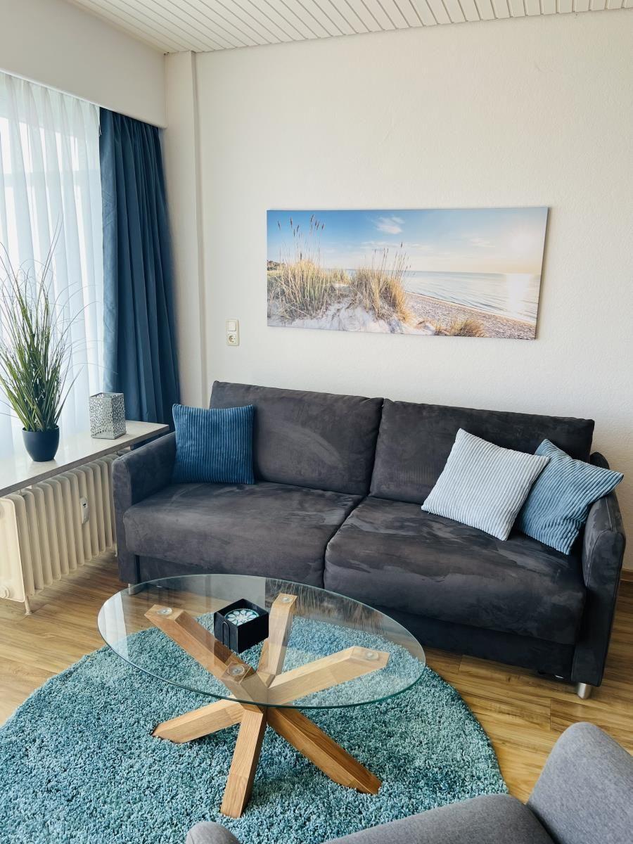 Ein Wohnzimmer mit grauem Sofa, Glas-Tisch und Strandbild an der Wand.