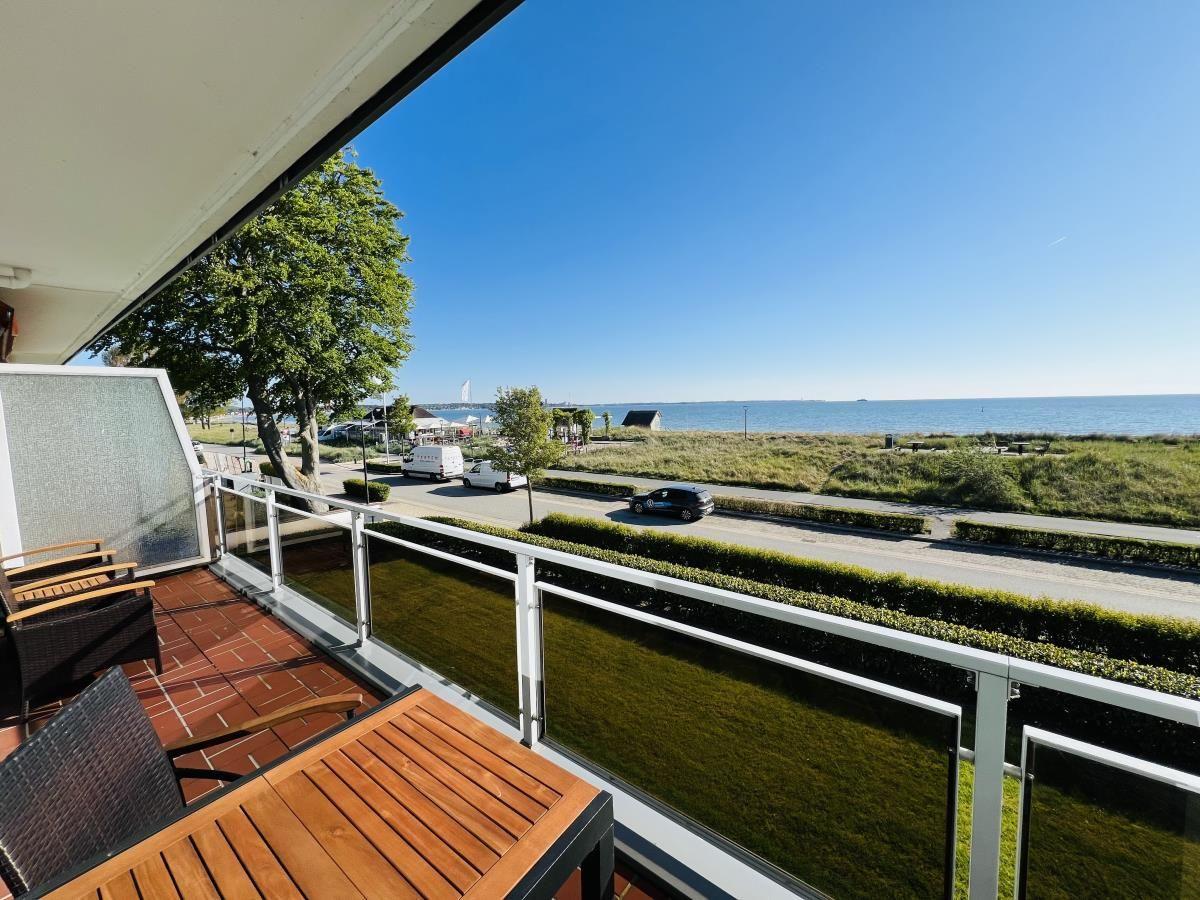 Terrasse mit Tisch und Stühlen, Blick auf Straße, Grünfläche und Meer.