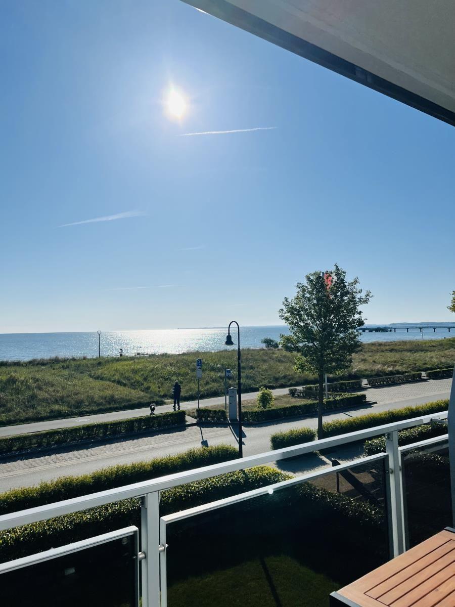 Balkon mit Blick auf Meer, Grünfläche und Promenade unter blauem Himmel.