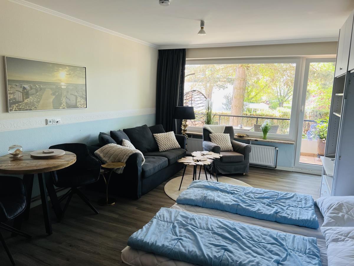 Wohnraum mit Schlafbereich, Sofa, Sessel und Balkon mit Blick auf Grünfläche.