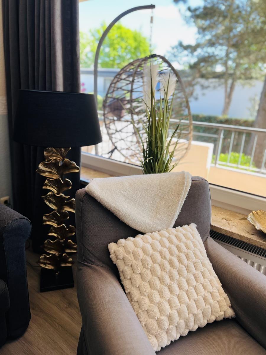 Ein grauer Sessel mit Kissen und Decke vor großem Fenster mit Blick auf Balkon.