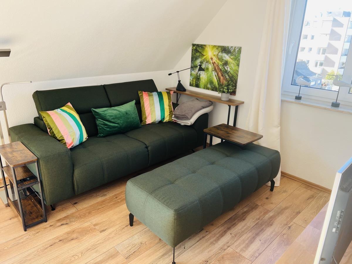 Ein Wohnzimmer mit grünem Sofa, Kissen und Holzboden. An der Wand hängt ein Bild.