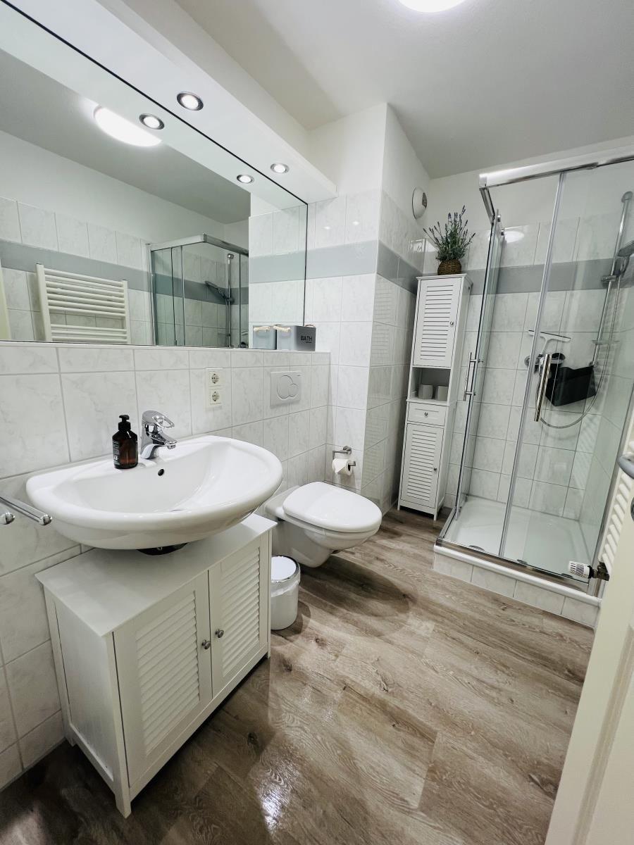 Badezimmer mit Dusche, Waschbecken und Toilette. Fliesenwand und Holzboden.