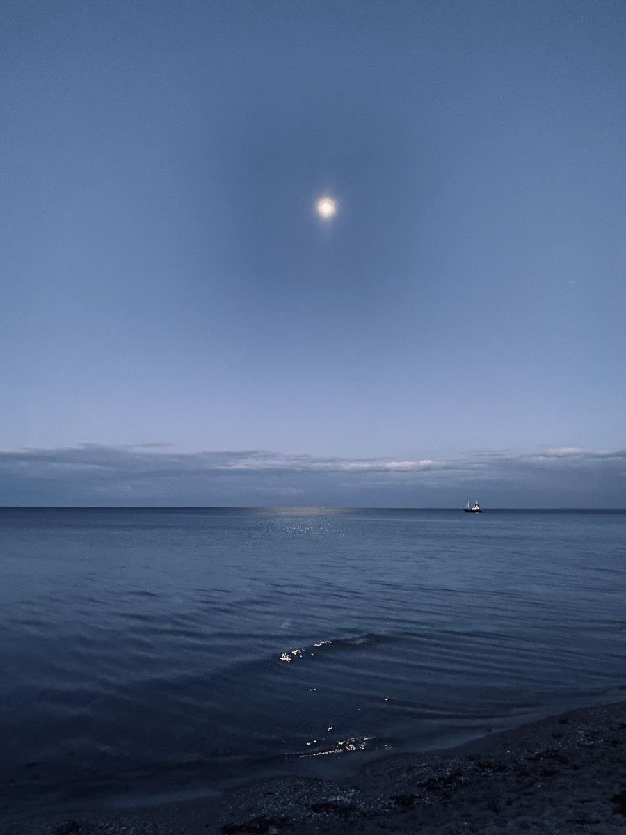 Moon over calm sea, small boat in background, beach in foreground.