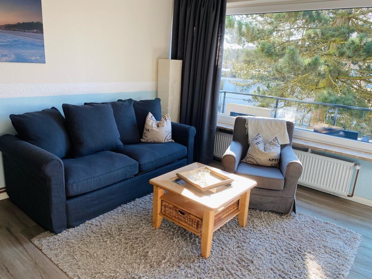 Wohnzimmer mit Sofa, Sessel und Couchtisch vor Fenster mit Blick auf Bäume.