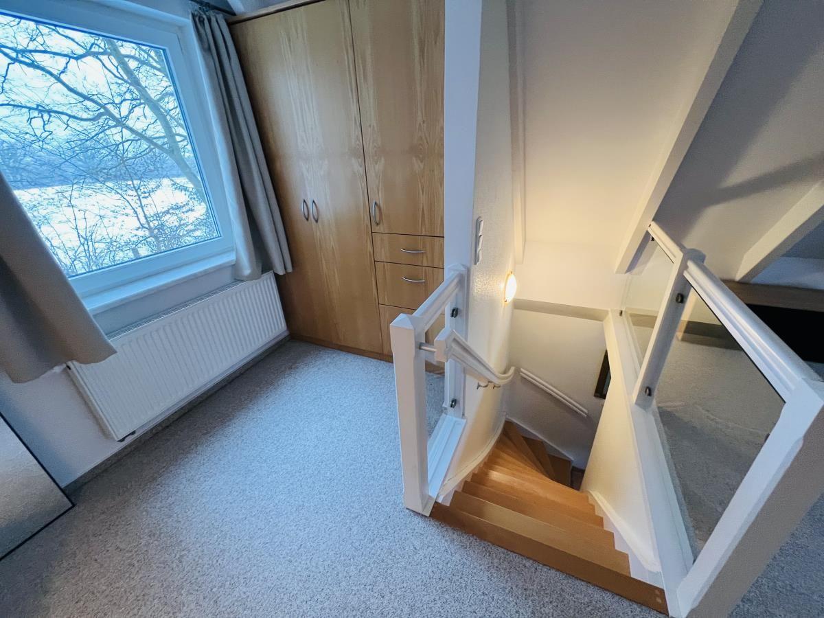 Treppenhaus mit Holztreppe, Garderobe und Fenster mit Blick auf Bäume.