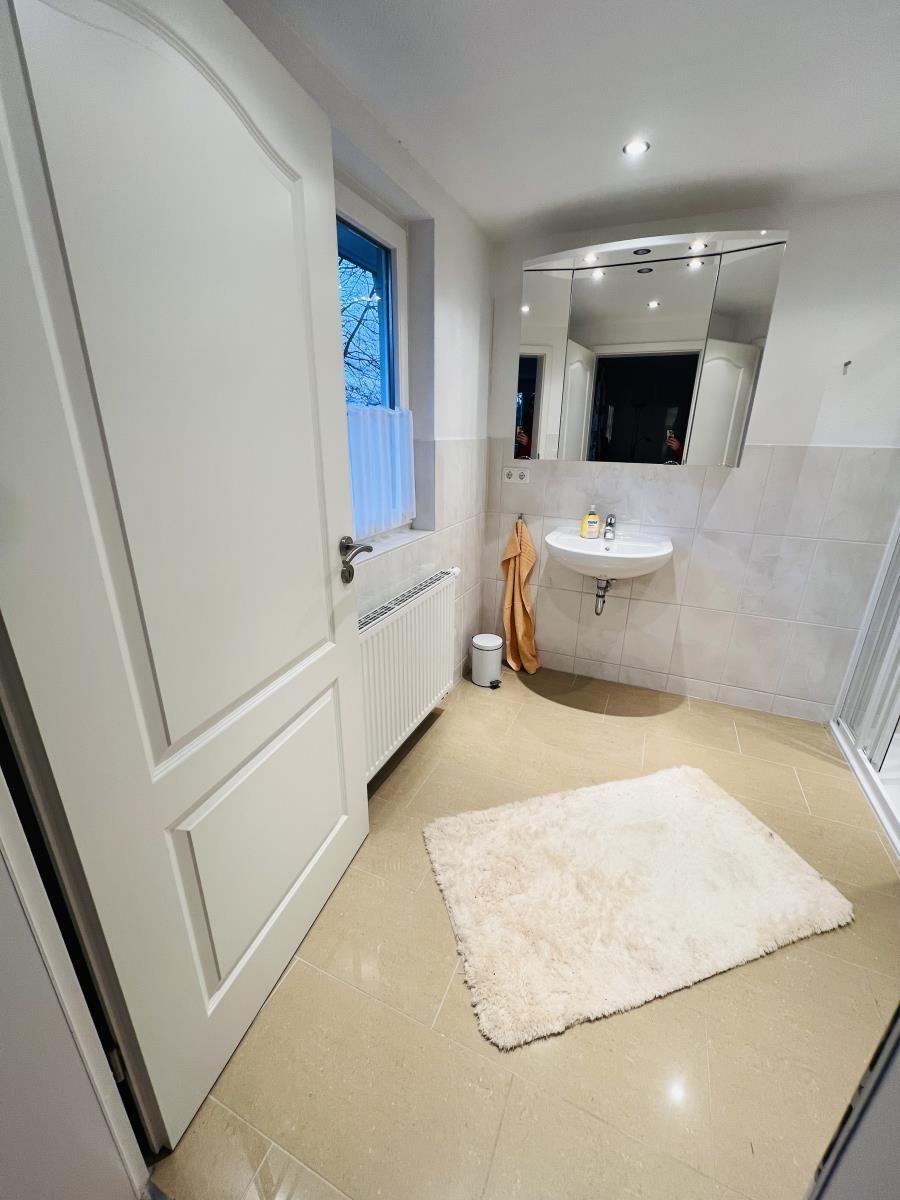 Badezimmer mit Waschbecken, Spiegel und Dusche. Fenster mit Vorhang und Teppich auf dem Boden.