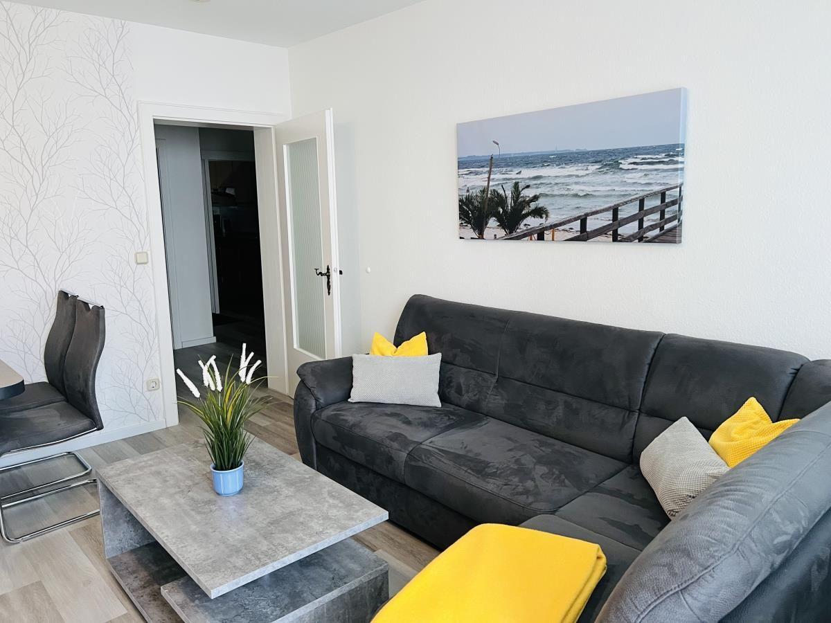 Wohnzimmer mit grauem Sofa, gelben Kissen und Küchenbereich im Hintergrund.