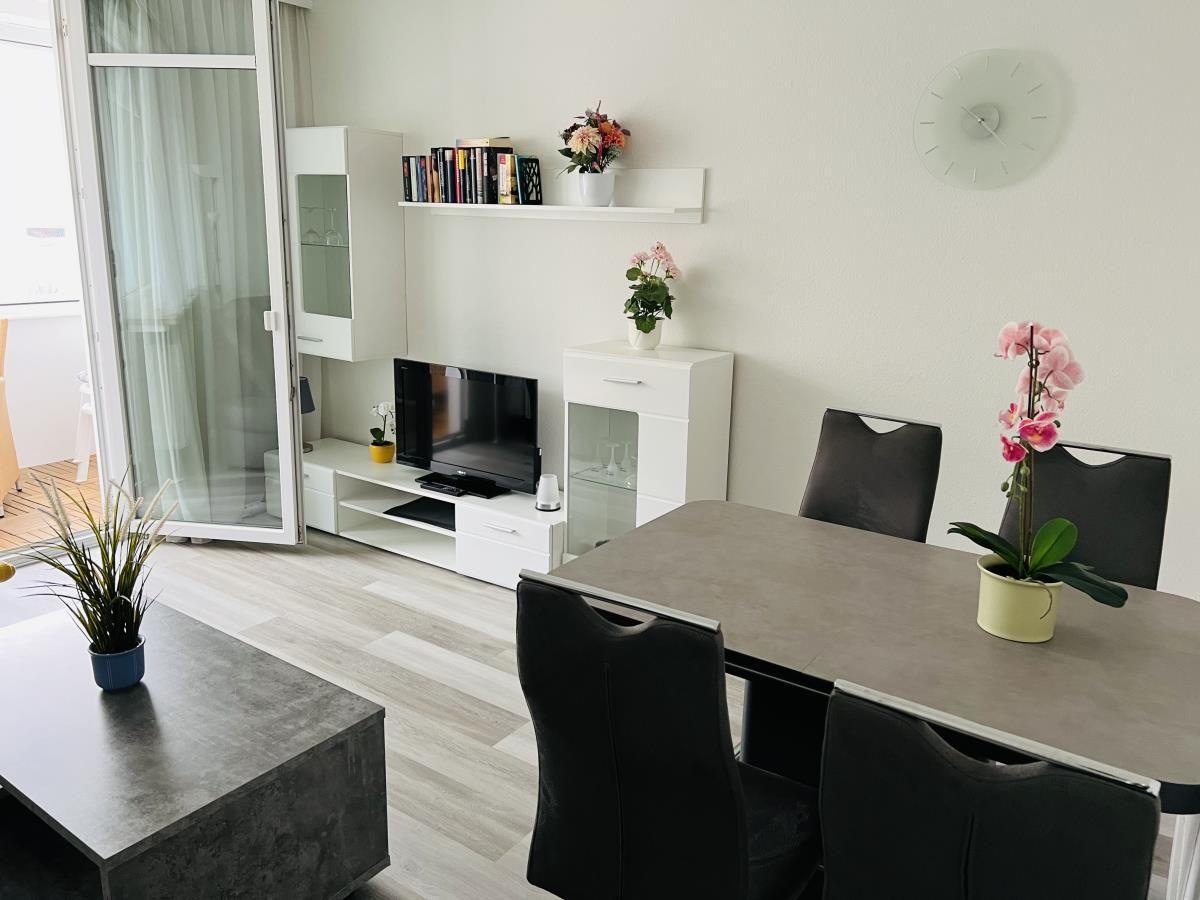 Wohn- und Essbereich mit TV, Tisch, Stühlen und Pflanzen. Terrasse über Glastür zugänglich.