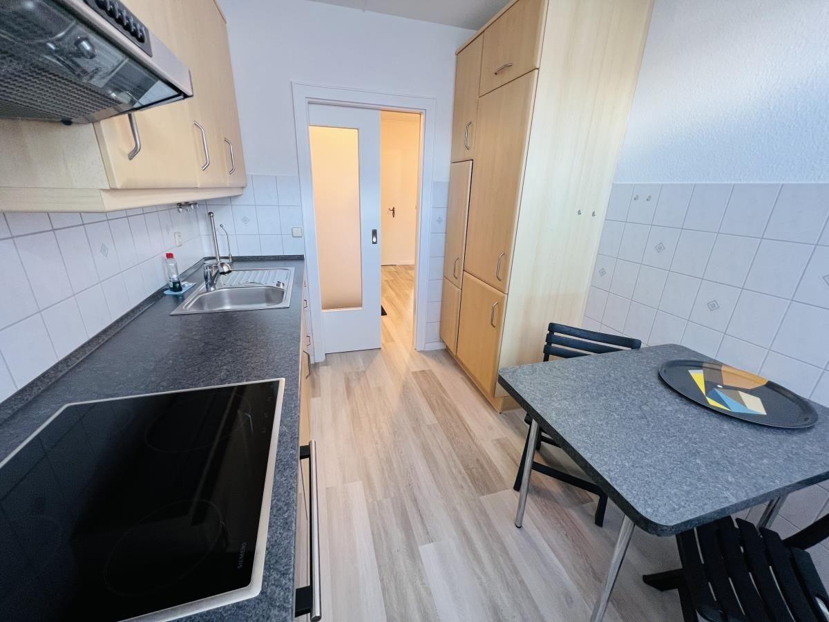 Küche mit Arbeitsplatte, Spüle und Kochfeld. Tisch mit Stühlen und Schrank im Hintergrund.