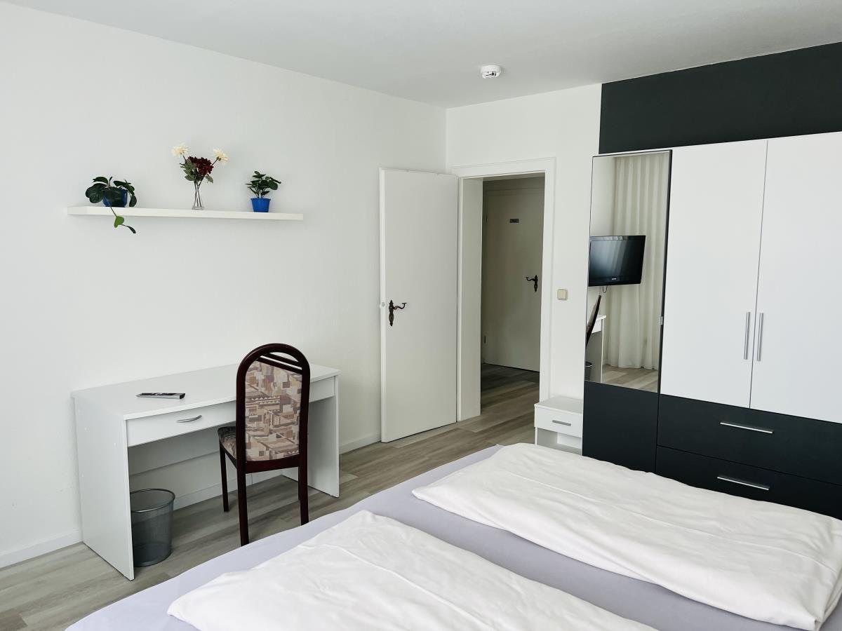 Schlafzimmer mit Bett, Schreibtisch, Kleiderschrank und Wandregal mit Pflanzen.