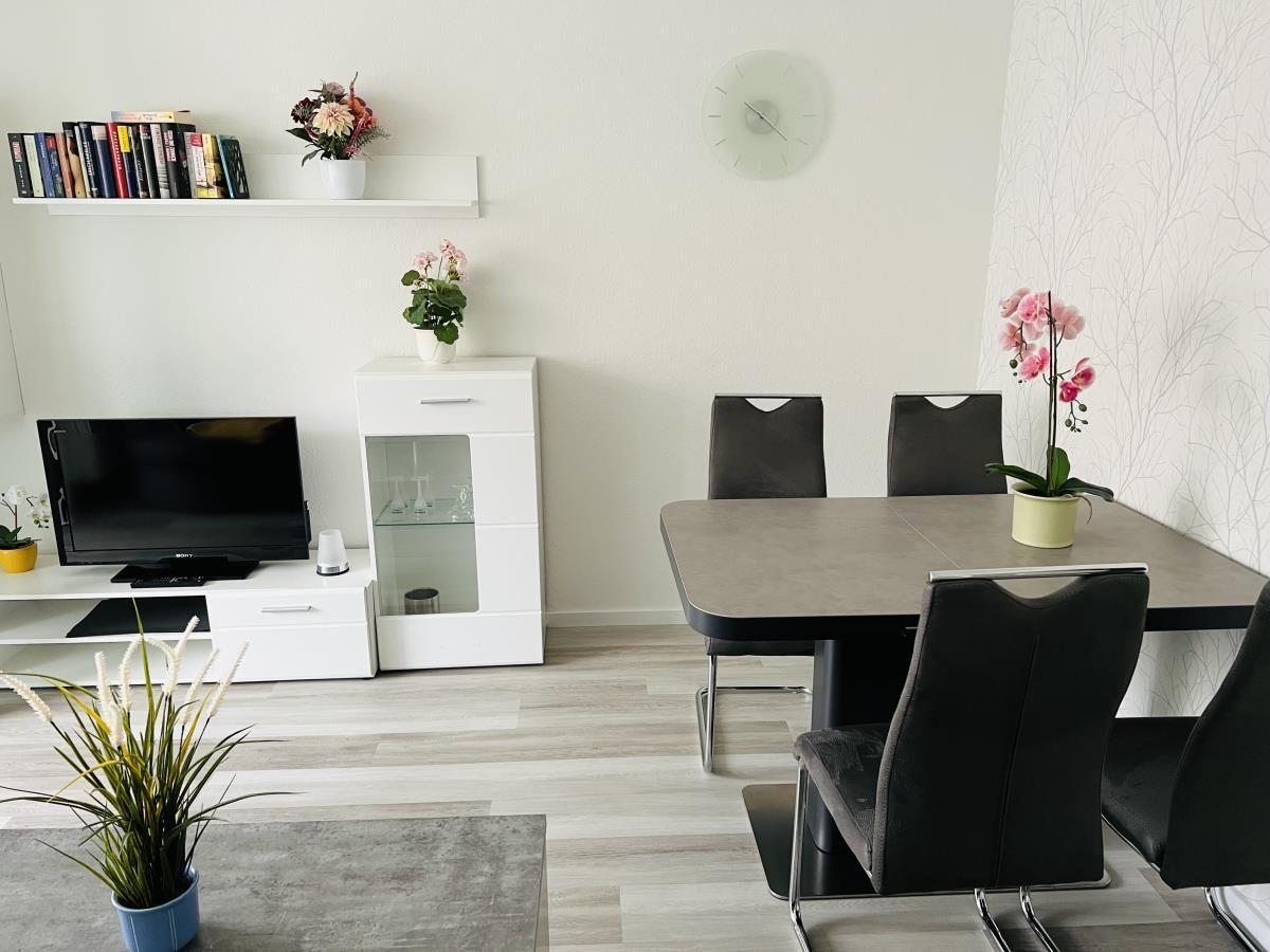 Esszimmer mit Tisch, Stühlen und Fernseher. Wanduhr und Pflanzen sind sichtbar.