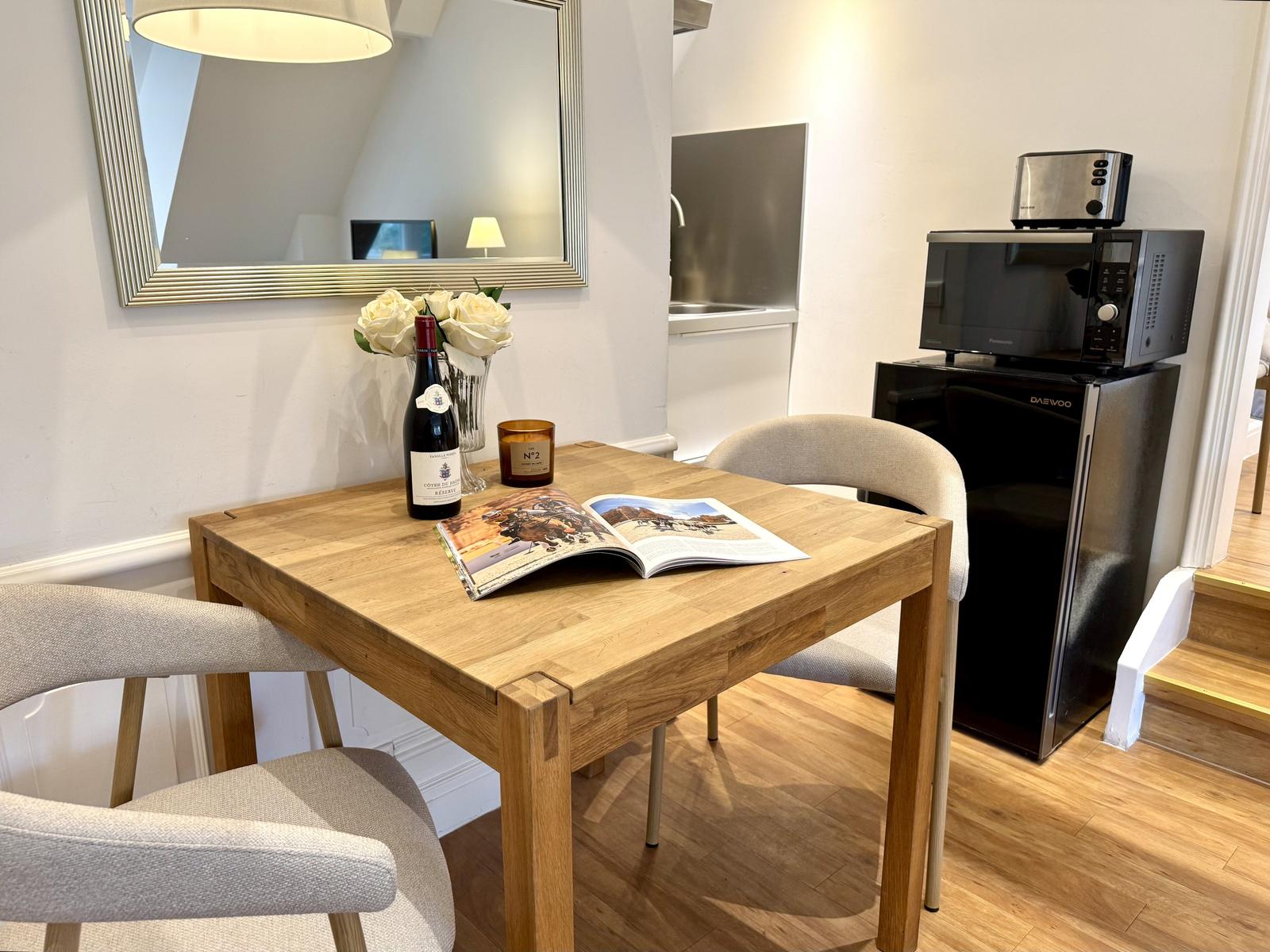 Holztisch mit Stühlen, Weinflasche und Zeitschrift. Küchenbereich mit Kühlschrank und Mikrowelle.