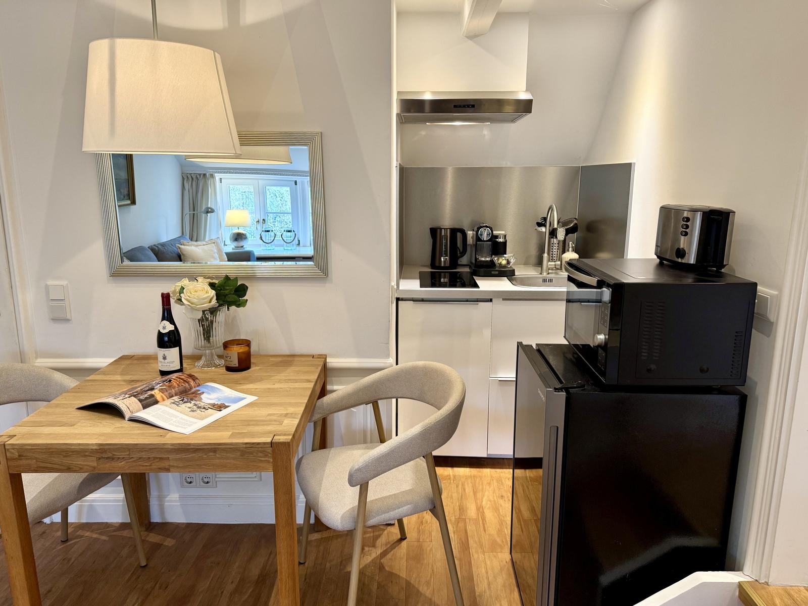 Essbereich mit Tisch, Stühlen und Spiegel. Küche mit Kühlschrank, Mikrowelle und Kochfeld.