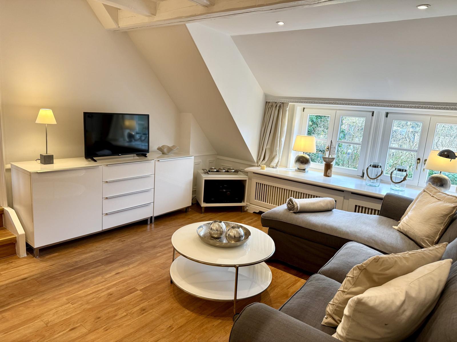 Wohnzimmer mit Sofa, TV und Fenster. Holzboden und weiße Möbel.