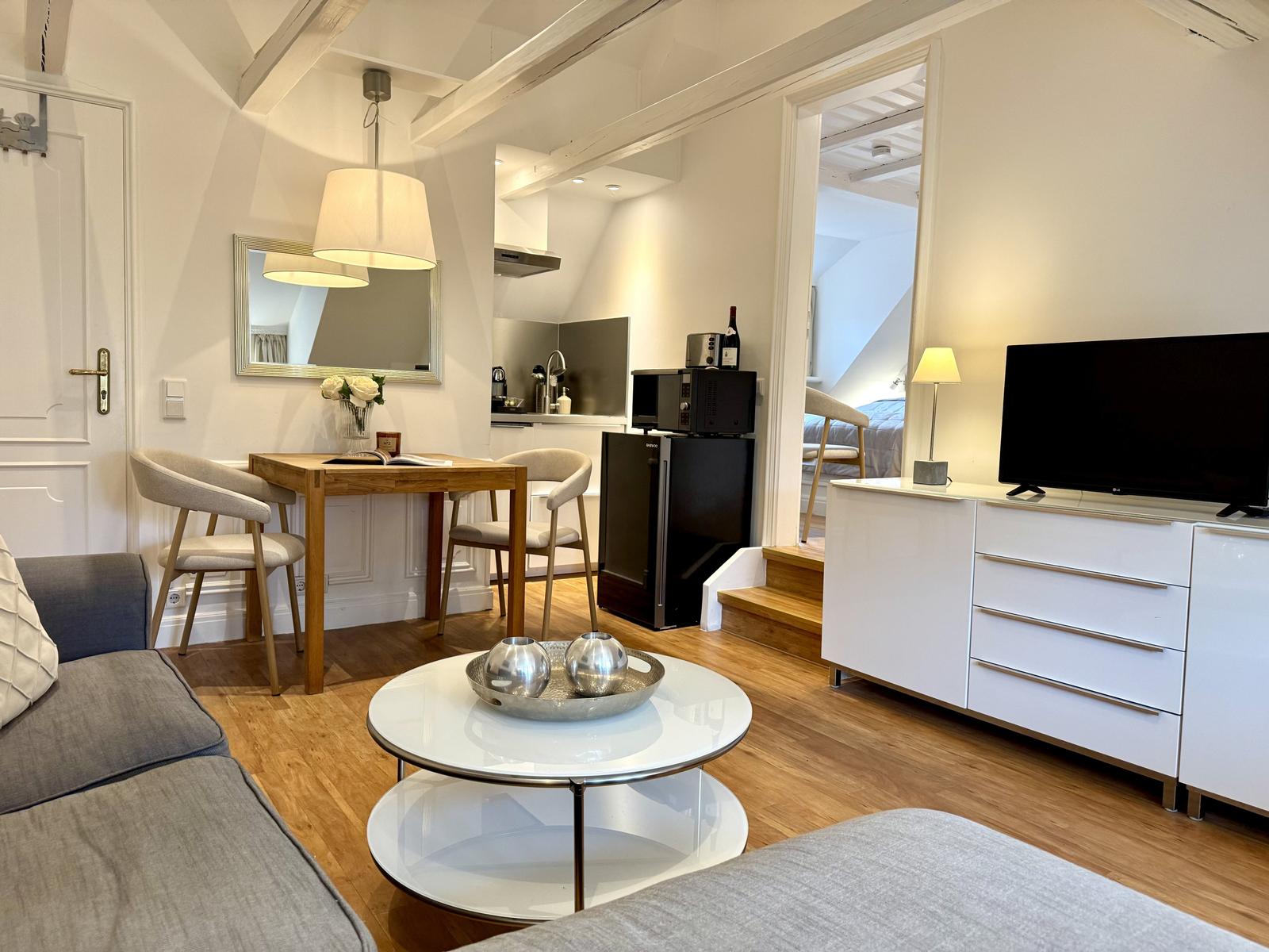 Wohnbereich mit Essplatz, Küchenzeile und TV-Schrank. Holzboden und weiße Wände.