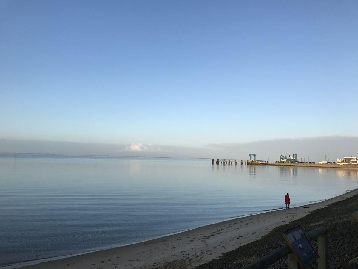 Ein Mensch in roter Jacke steht am Strand neben einem Steg. Im Hintergrund sind Gebäude und Wasser zu sehen.