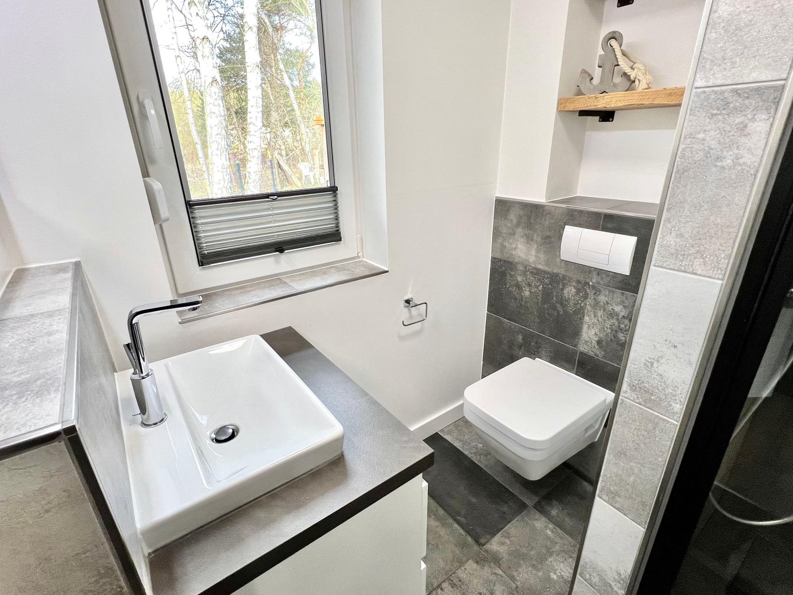 Badezimmer mit Waschbecken, WC und Dusche. Fenster mit Blick auf Bäume.