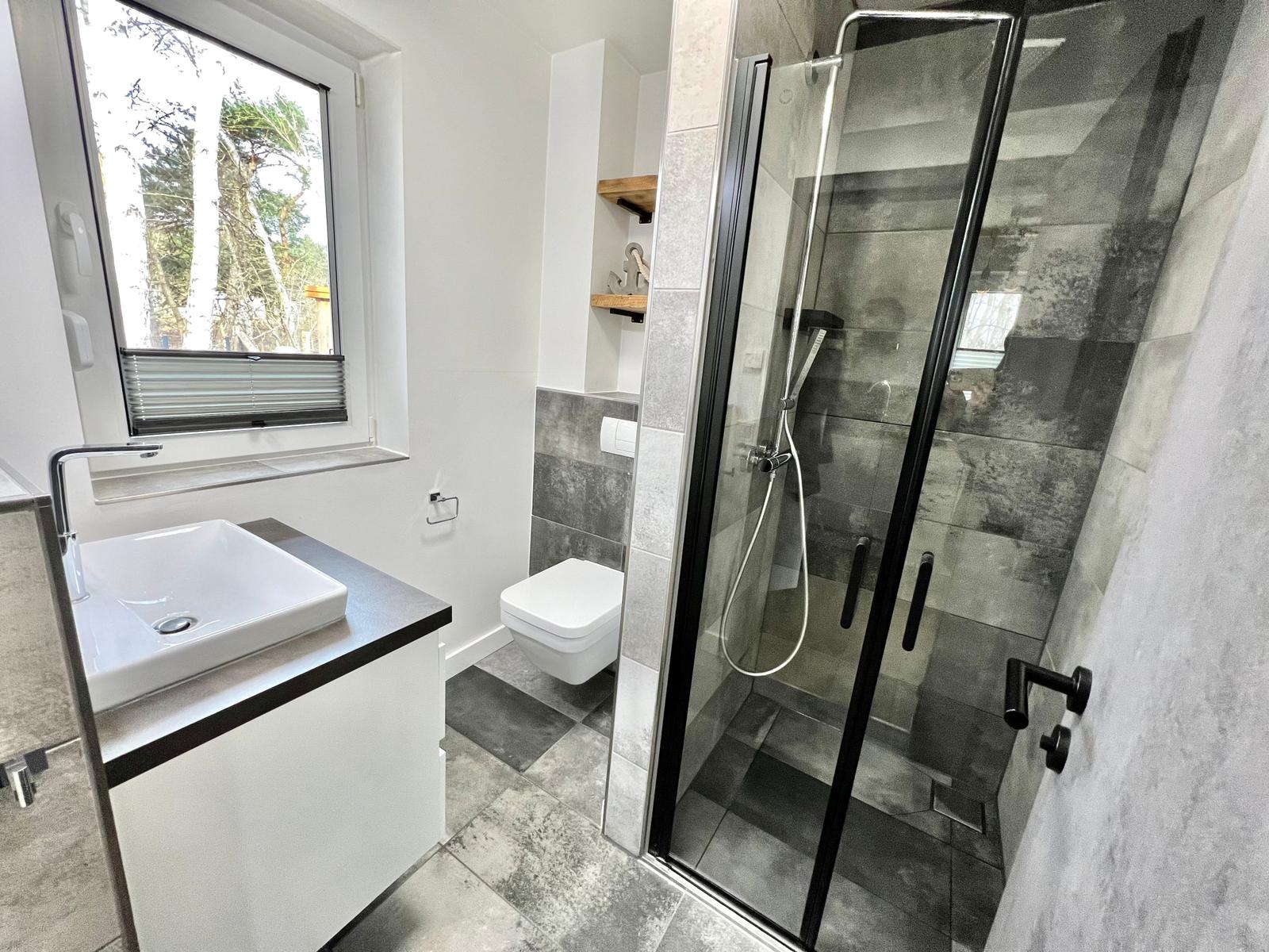 Badezimmer mit Dusche, Waschbecken und Toilette. Fenster mit Blick auf Bäume.