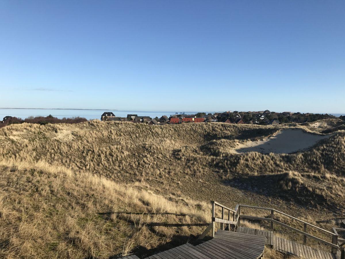 Holzsteg führt durch Dünen zu einem Stranddorf mit roten Häusern und Blick aufs Meer.