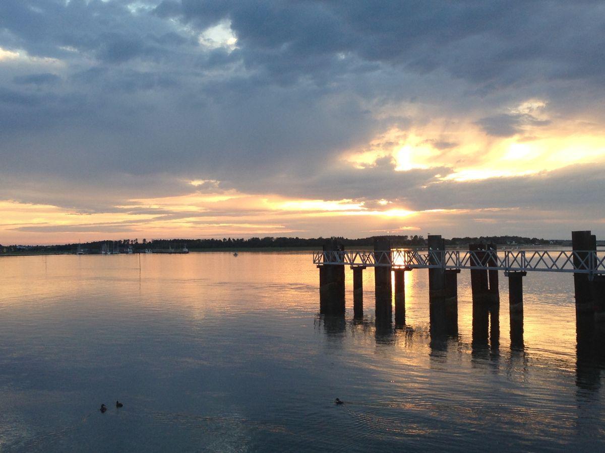 Sonnenuntergang über dem Wasser mit Pier und Booten im Hintergrund.