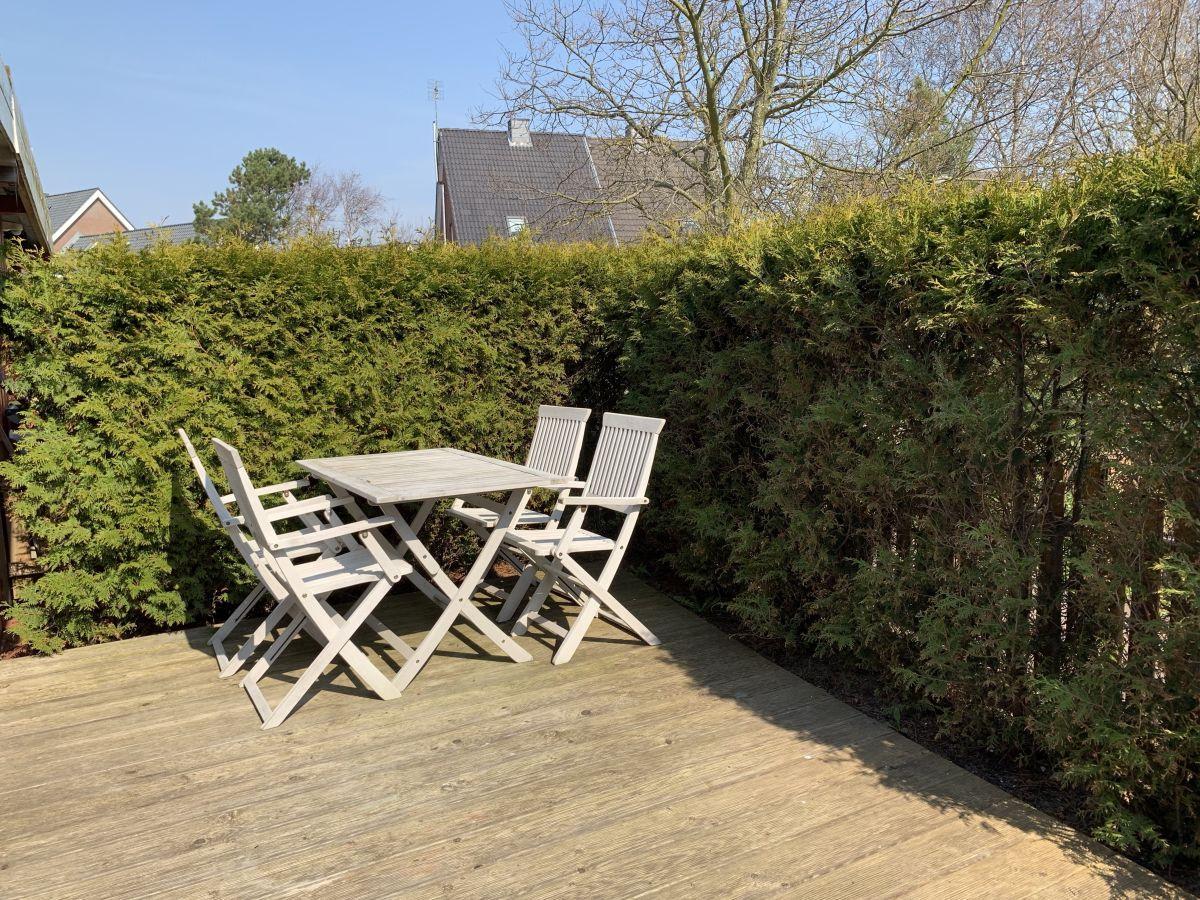 Holzterrasse mit Tisch und vier Stühlen, umgeben von grünen Hecken.