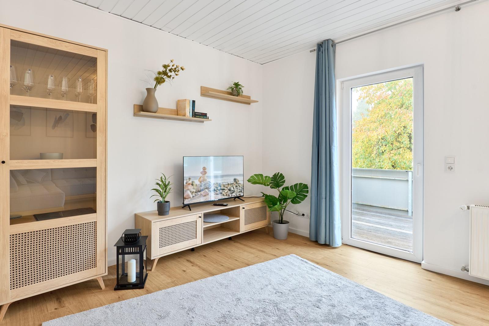 Wohnzimmer mit TV, Schrank und Balkontür. Holzboden und Pflanzen.