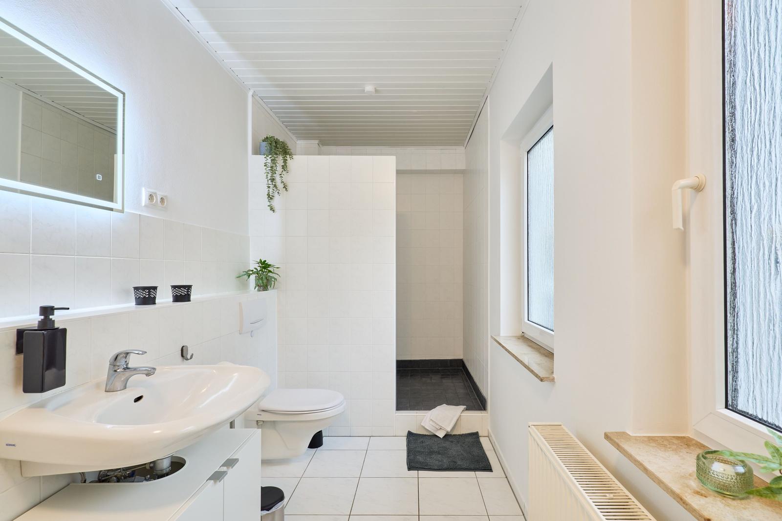 Badezimmer mit Dusche, Toilette und Waschbecken. Fenster und Pflanzen sorgen für Licht.