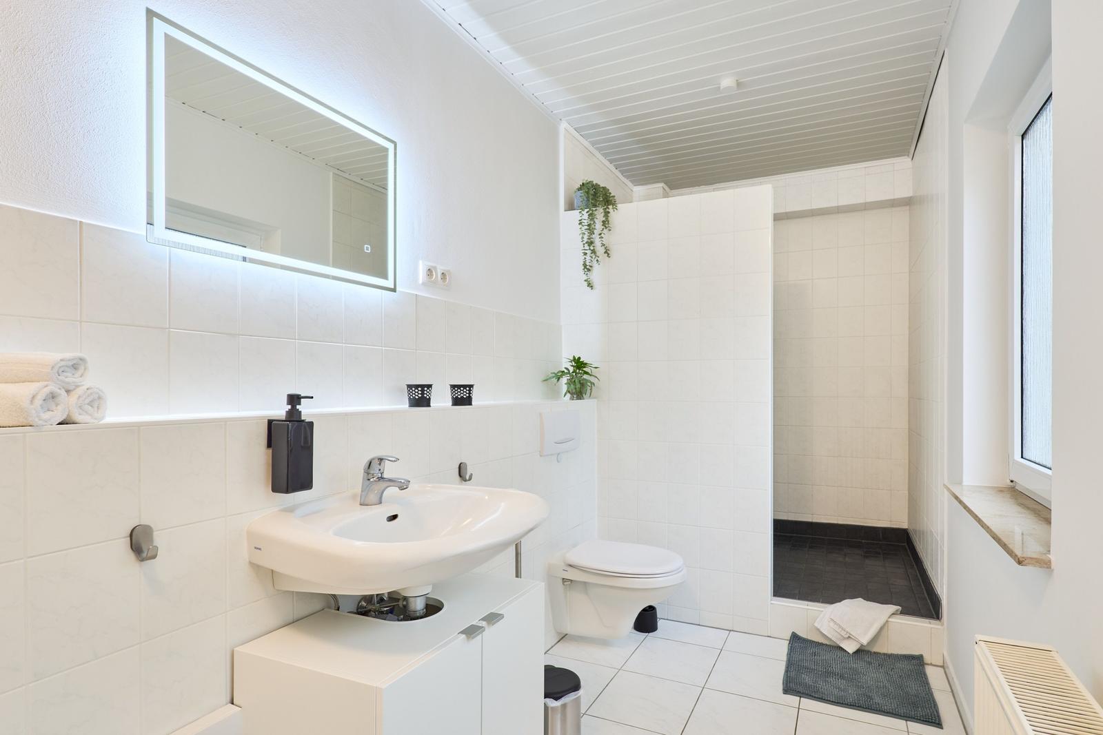 Badezimmer mit Waschbecken, Toilette und Dusche. Weiße Fliesen und beleuchtetes Spiegel.