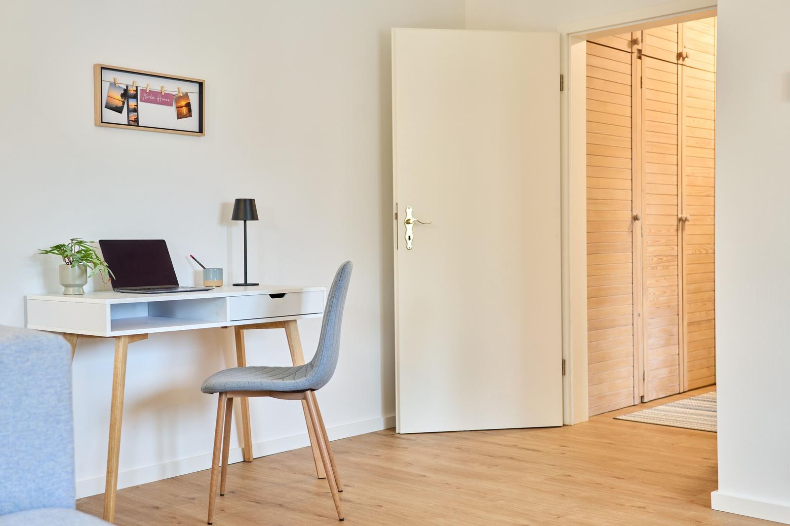 Arbeitsplatz mit Schreibtisch, Stuhl und Laptop. Tür führt in Schlafzimmer mit Holzschrank.