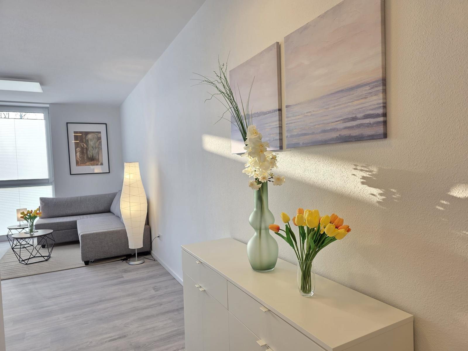 Wohnbereich mit Sofa, Beistelltisch und Stehlampe. Anrichte mit Blumen und Wandbildern.