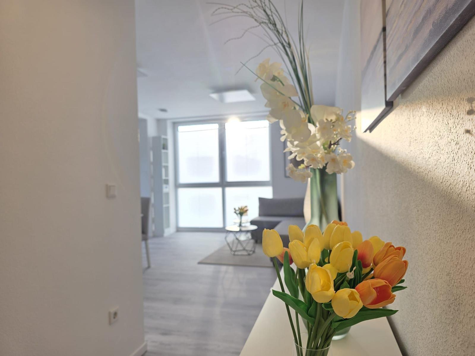 Vorraum mit Blumen, Blick in Wohnbereich mit Sofa und Fenster.