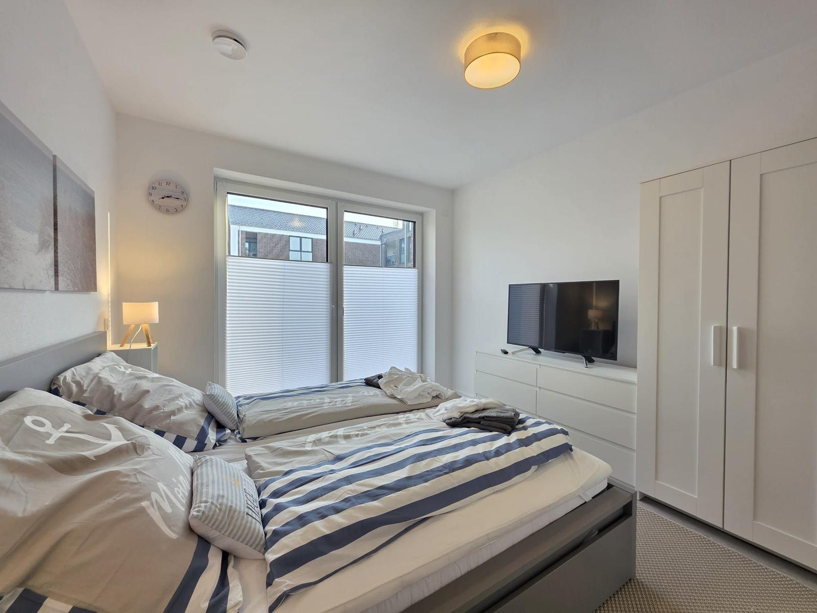 Schlafzimmer mit Doppelbett, TV und Kleiderschrank. Fenster mit Jalousien.