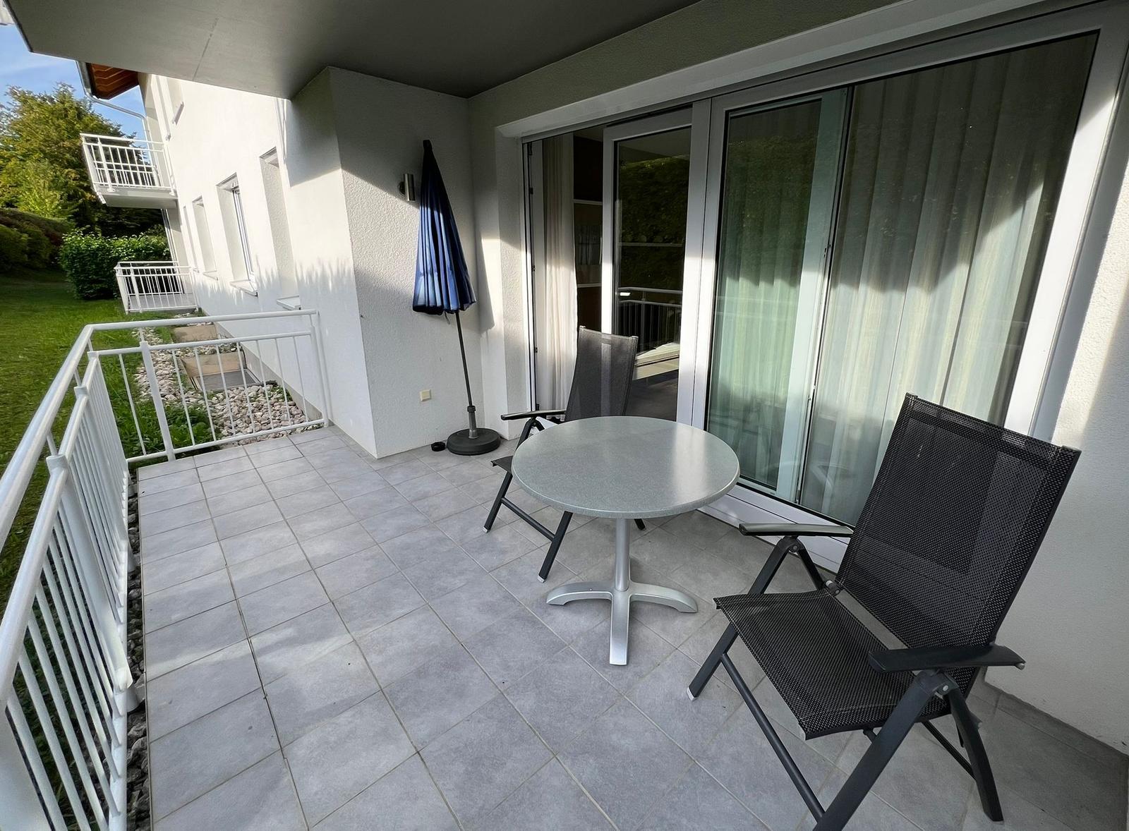 Terrace with table, two chairs, and blue umbrella. Glass door leads into the house.