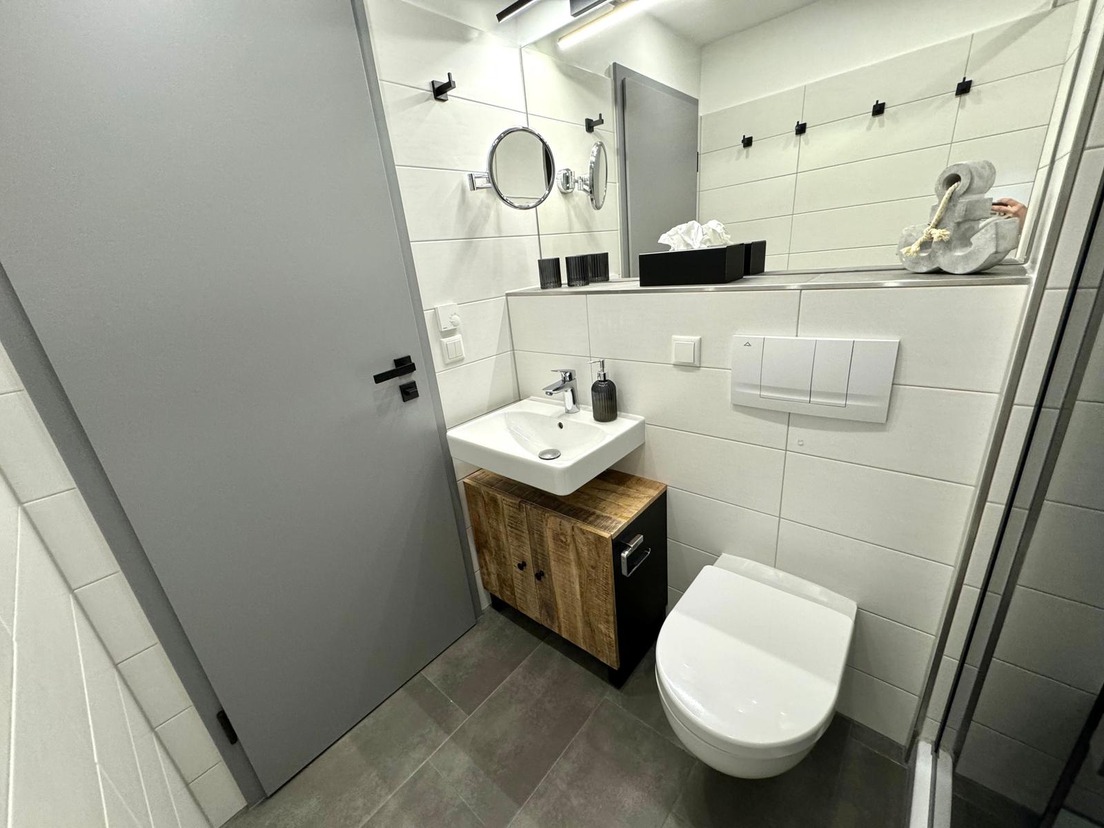 Badezimmer mit Waschbecken, Toilette und Spiegel. Holzunterschrank und graue Fliesen.
