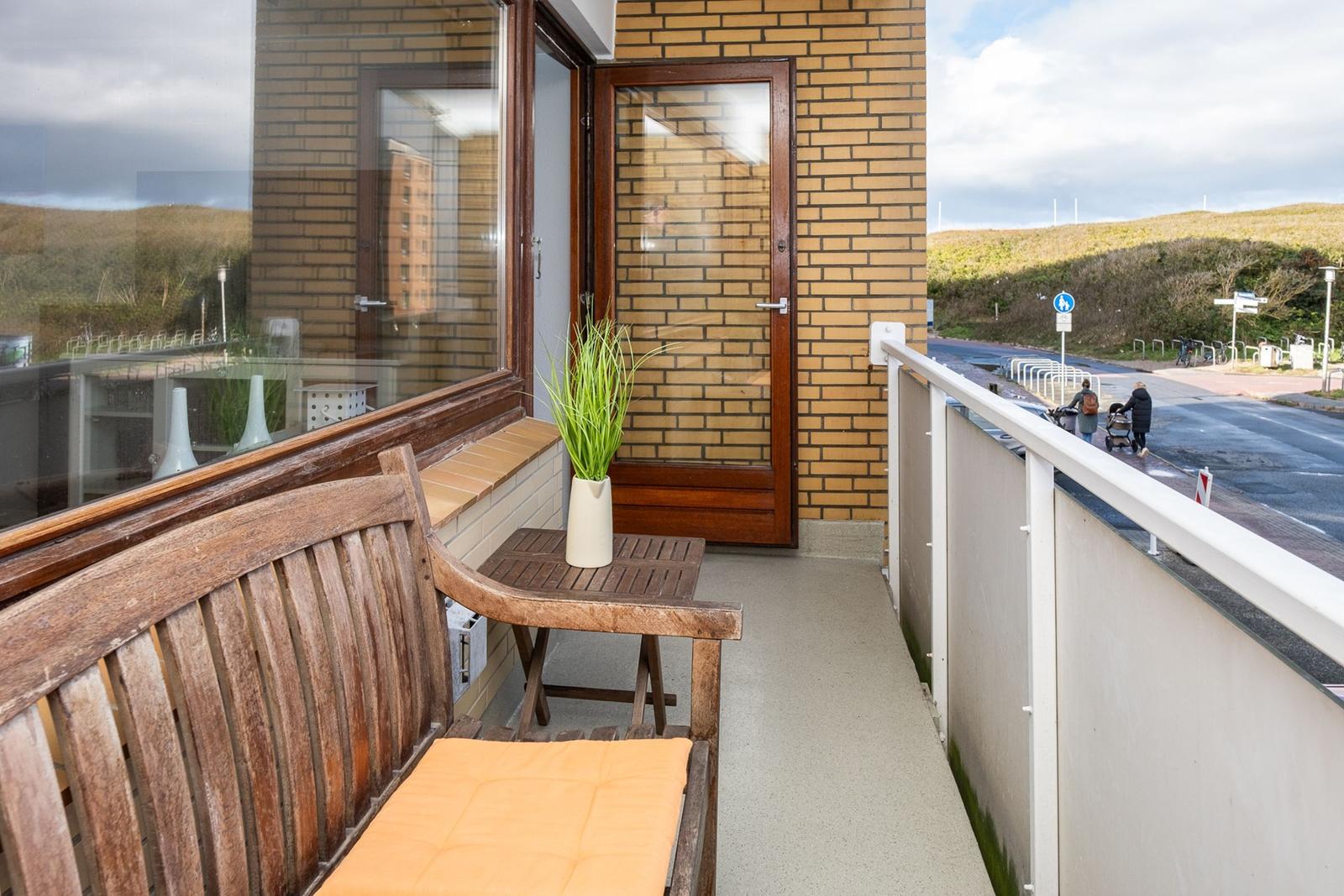 Balcony with wooden bench, table, and plant. View of street and hills.