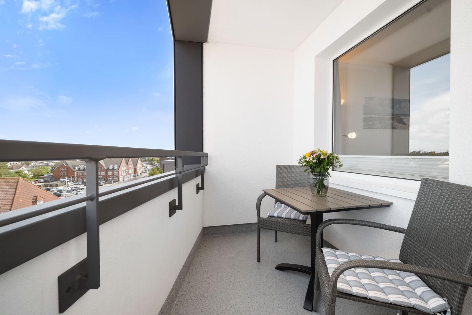 Balkon mit Tisch, zwei Stühlen und Blumenarrangement. Blick auf Wohngebiet unter blauem Himmel.