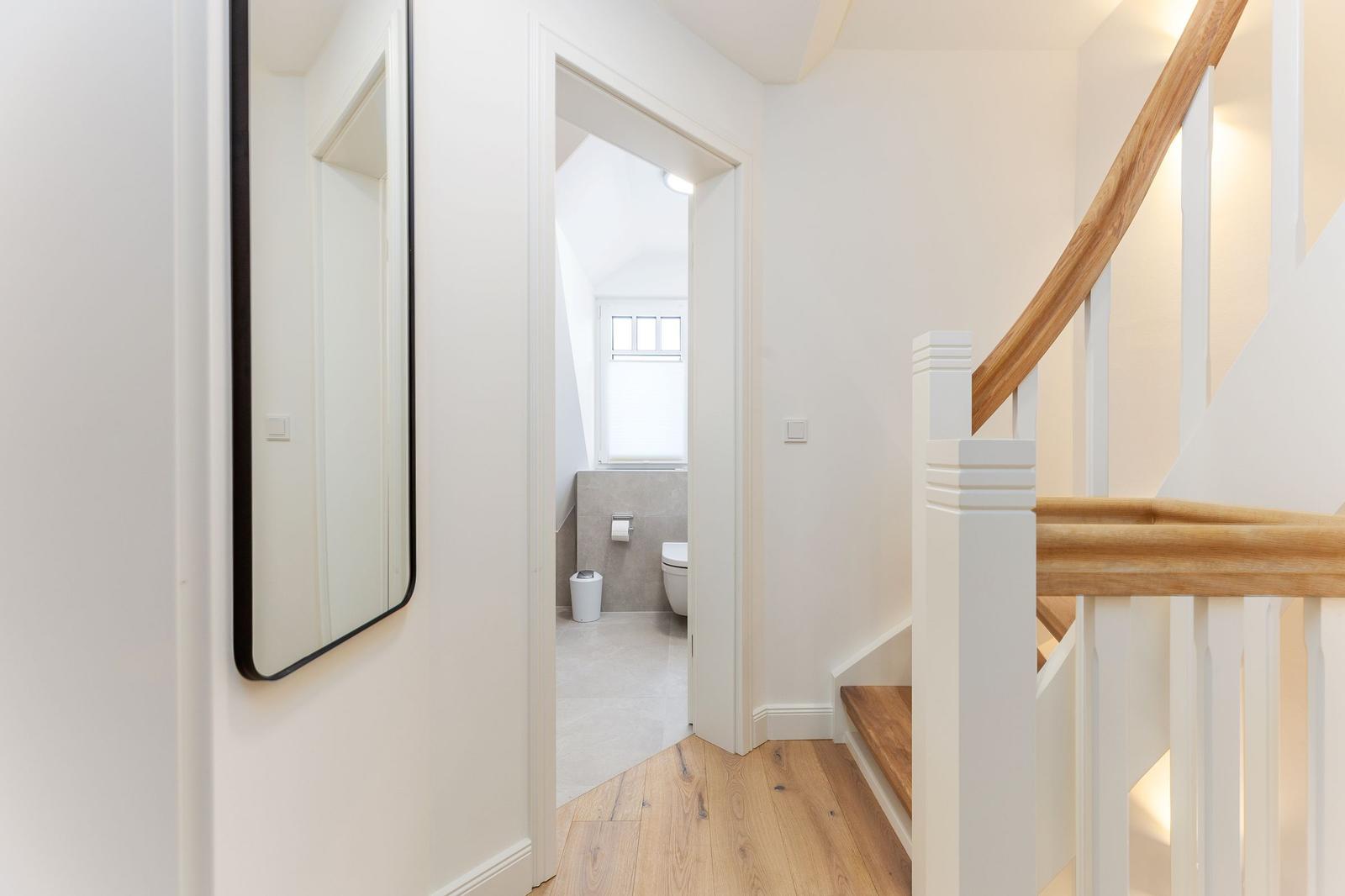 Haus mit Holztreppe und Spiegel. Im Hintergrund ist ein Badezimmer mit Toilette und Fenster.