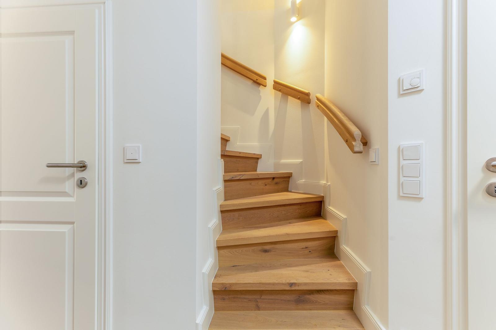 Holzgetreppe mit Geländer führt nach oben. Weiße Wände und Lichtschalter sind sichtbar.