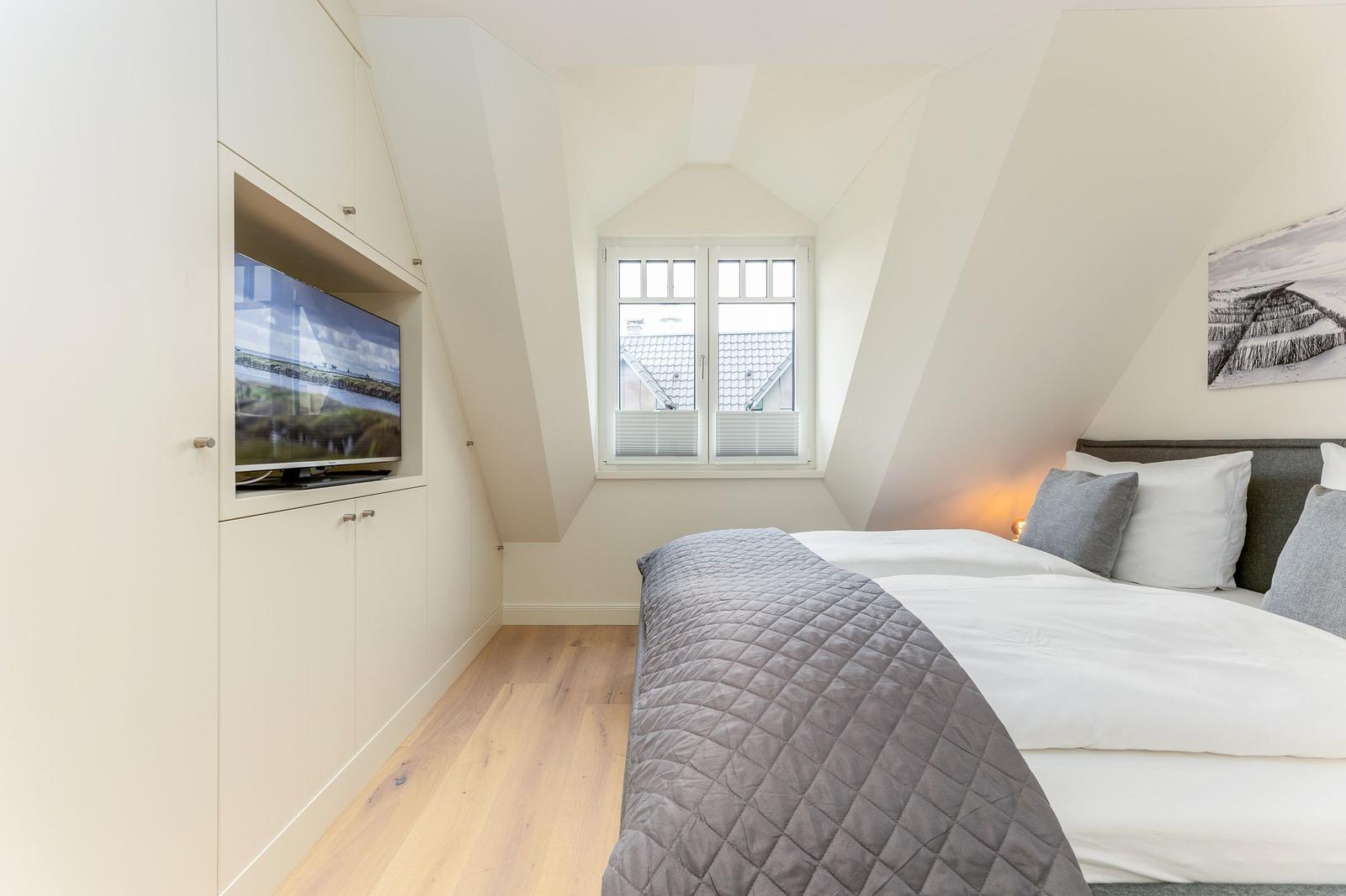 Schlafzimmer mit Dachschrägen, Bett, Fenster und eingebautem Fernseher.