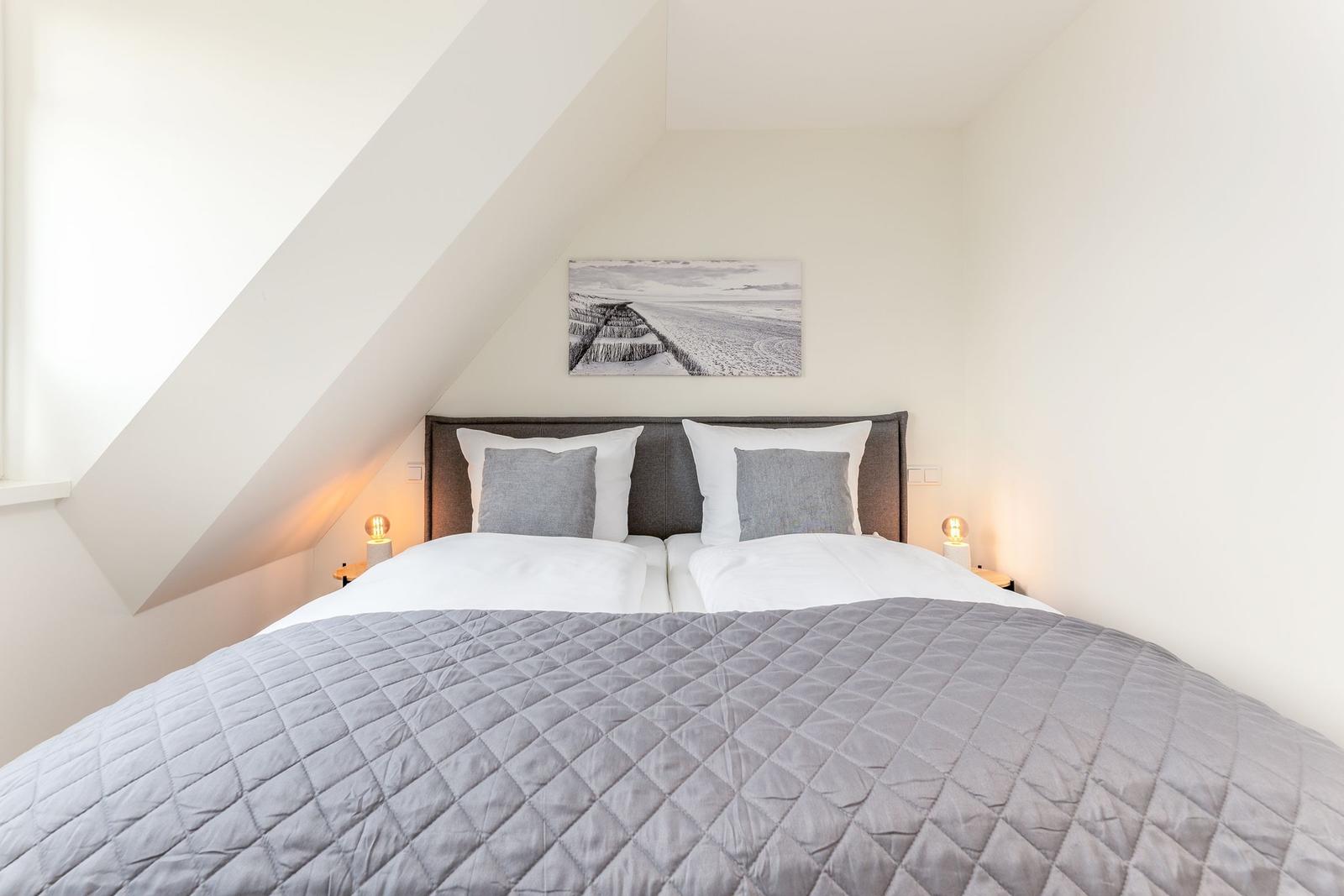 Doppelbett mit grauer Daunendecke unter schrägen Dachfenstern. Wandbild zeigt Strand.