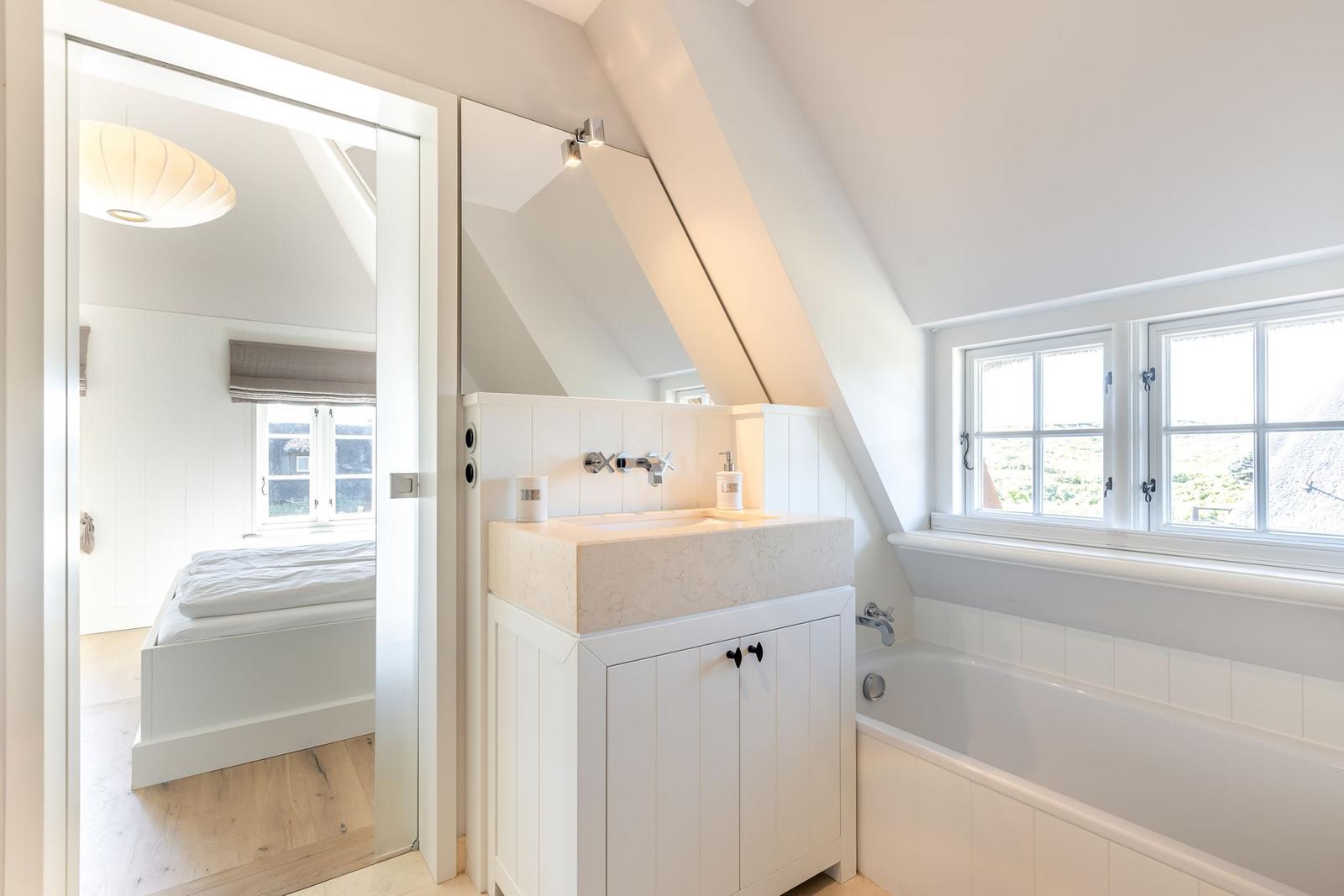 Badezimmer mit Badewanne und Waschbecken. Durchgang in Schlafzimmer mit Bett und Fenster.