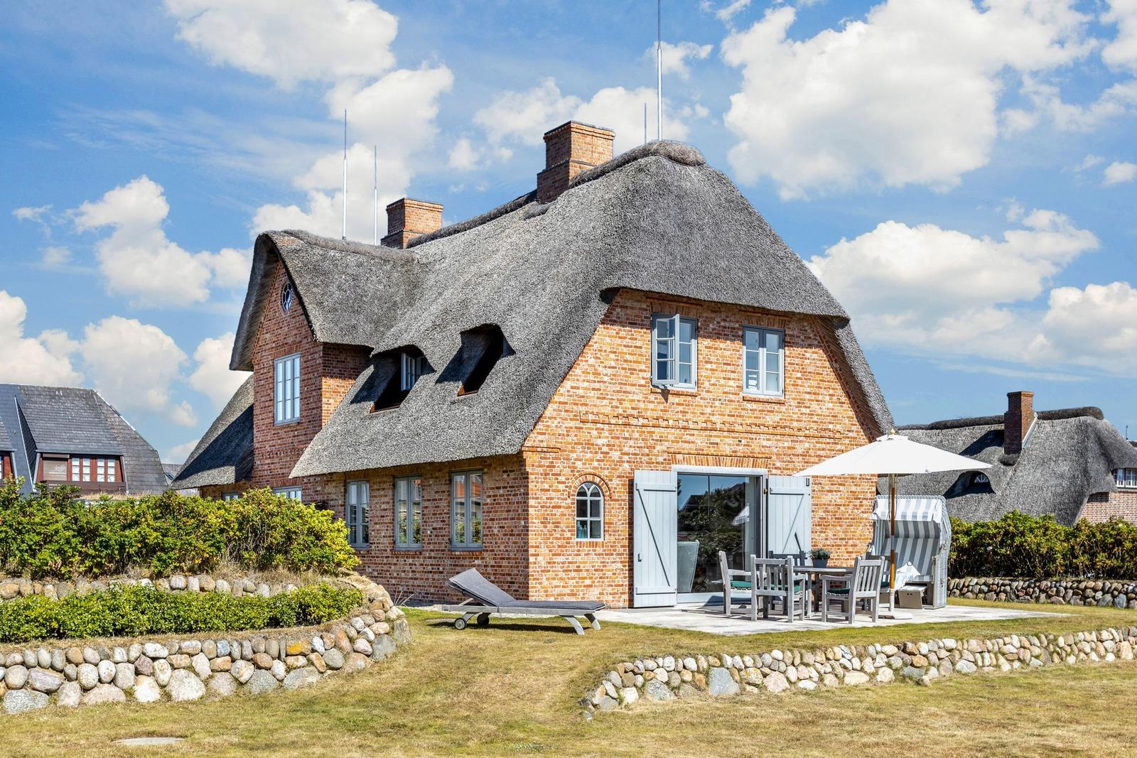 Ein Haus mit Strohdach, Terrasse und Garten. Ein Liegestuhl und Tisch sind draußen.