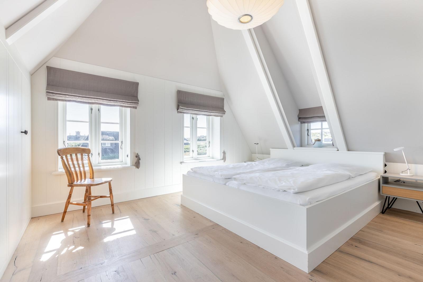 Schlafzimmer mit Dachschräge, weißem Bett, Holzboden und zwei Fenstern mit Jalousien.