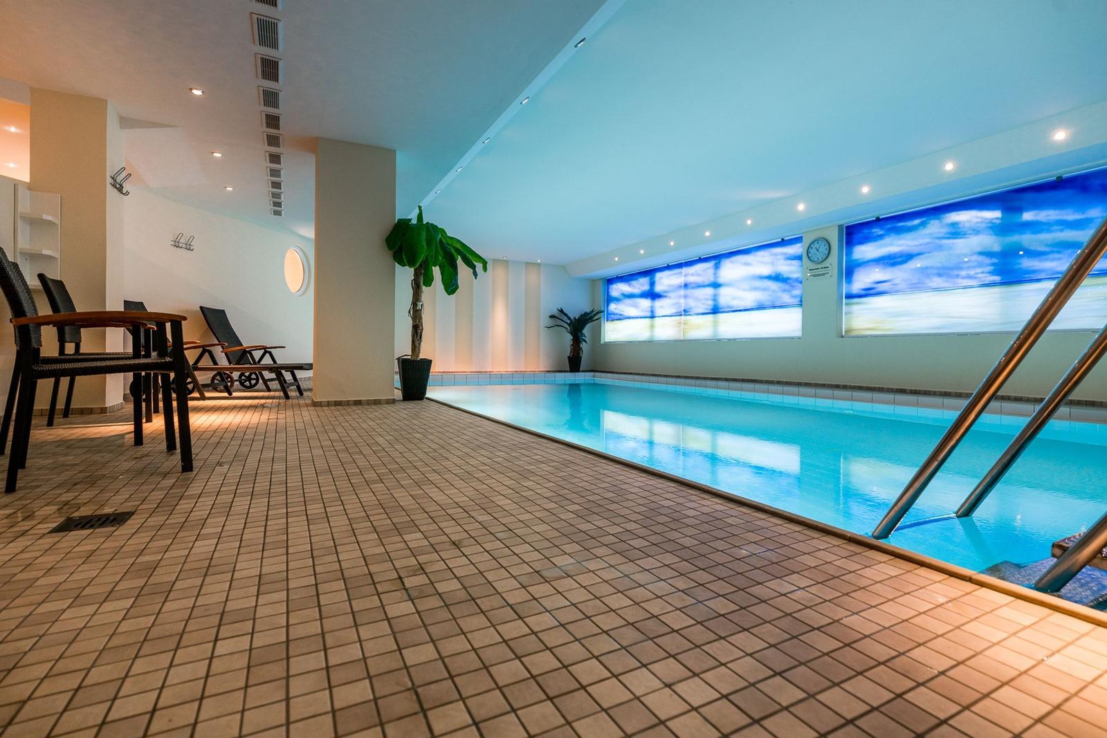 Indoorpool mit Liegestühlen und Pflanzen. Decke mit Beleuchtung. Wand mit Fensterbild.
