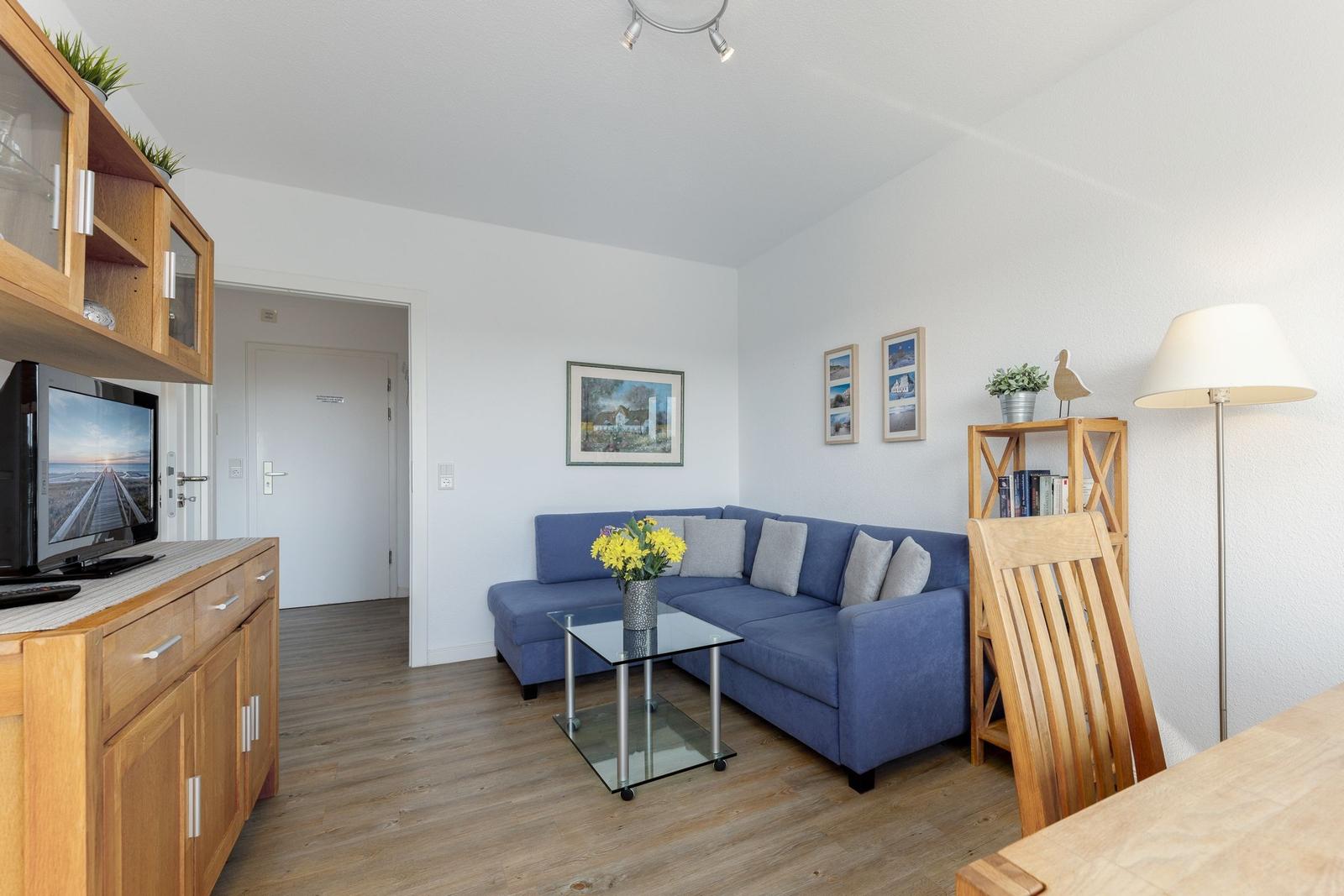 Wohnzimmer mit blauem Sofa, Glas-Tisch und Holzmöbeln. Fernseher und Bilder an der Wand.