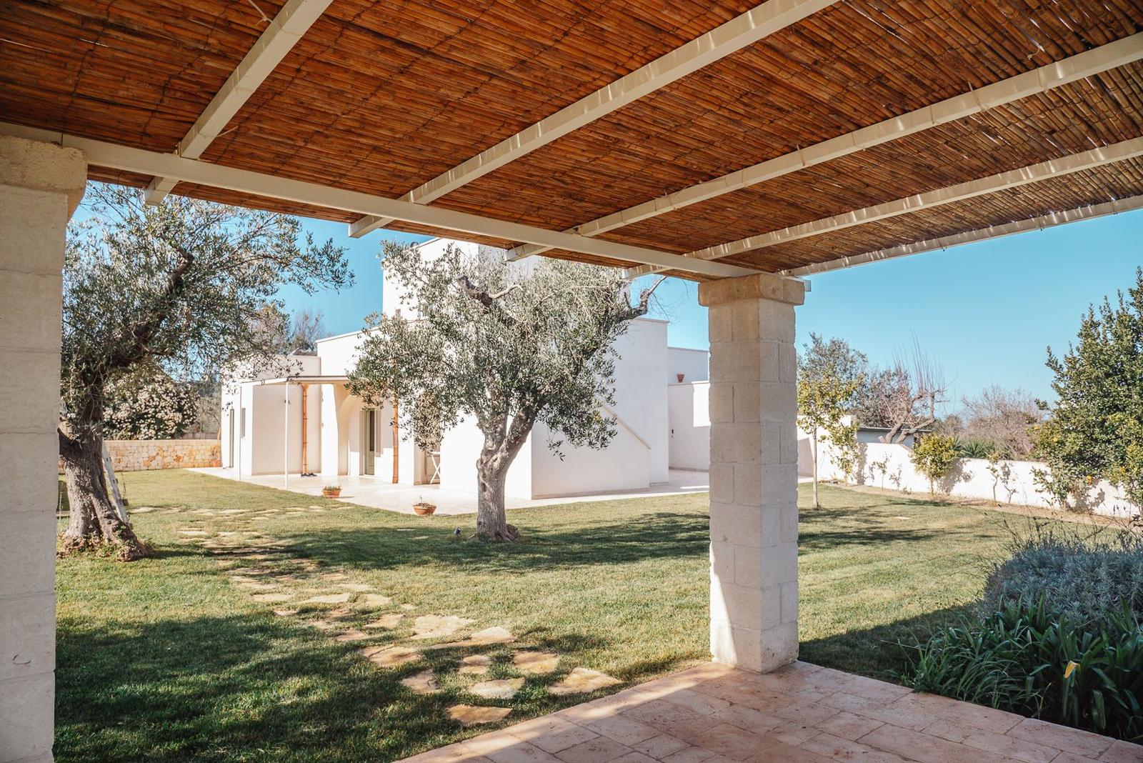 Patio with thatched roof overlooking white villa and olive trees.